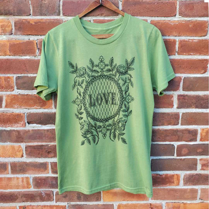Rare Breed Organic Apparel's light green organic cotton Love t-shirt hangs against a red brick wall.  This certified organic graphic tee features a black screen print of a vintage illustration containing the word "Love" within an ornate circle, and surrounded by roses, leaves and floral emblems.  Eco-friendly.  Ethically made.  And made with love.