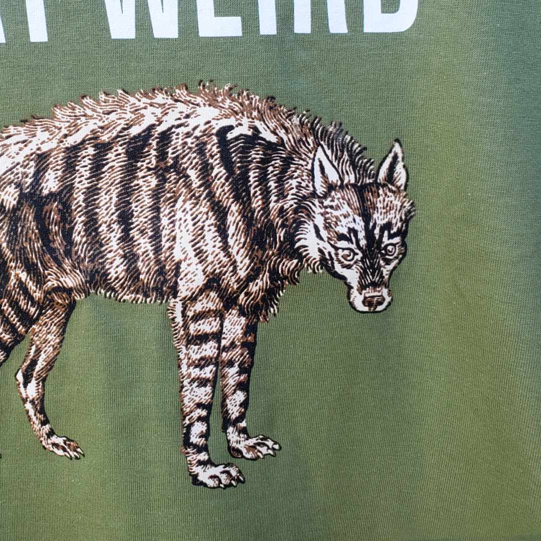 Closeup of Rare Breed Organic Apparel's organic cotton T-shirt featuring restored historical image of a sassy hyena staring down the camera and bold white characters proclaiming "STAY WEIRD" on an olive T-shirt.