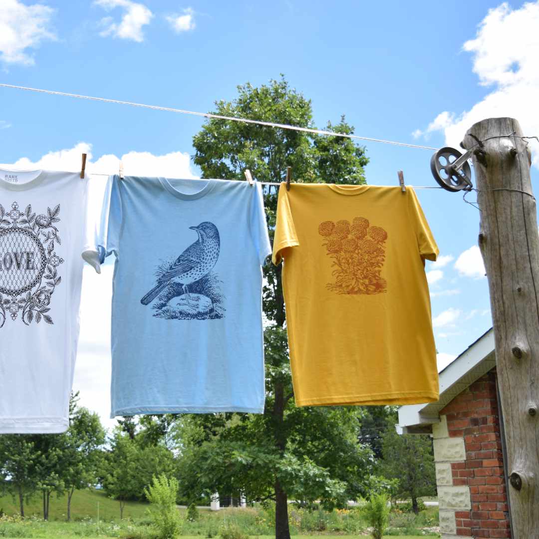 Three t-shirts by Rare Breed Organic Apparel hang on a rustic clothes line outside of a farm house. In the background are trees and a bright blue sky. The T-shirts show vintage illustrations from historical literature: A love vignette with roses on a white t-shirt, a navy blue bird on a light blue t-shirt and rust marigolds on a mustard t-shirt.