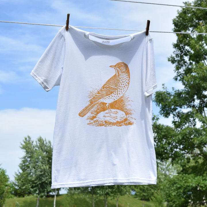 This white, 100% organic cotton Rare Breed t-shirt features a vintage wood cut image of a song bird.  The T-shirt hangs outdoors from a clothesline against of backdrop of natural trees, grass, and a bright blue sky.  