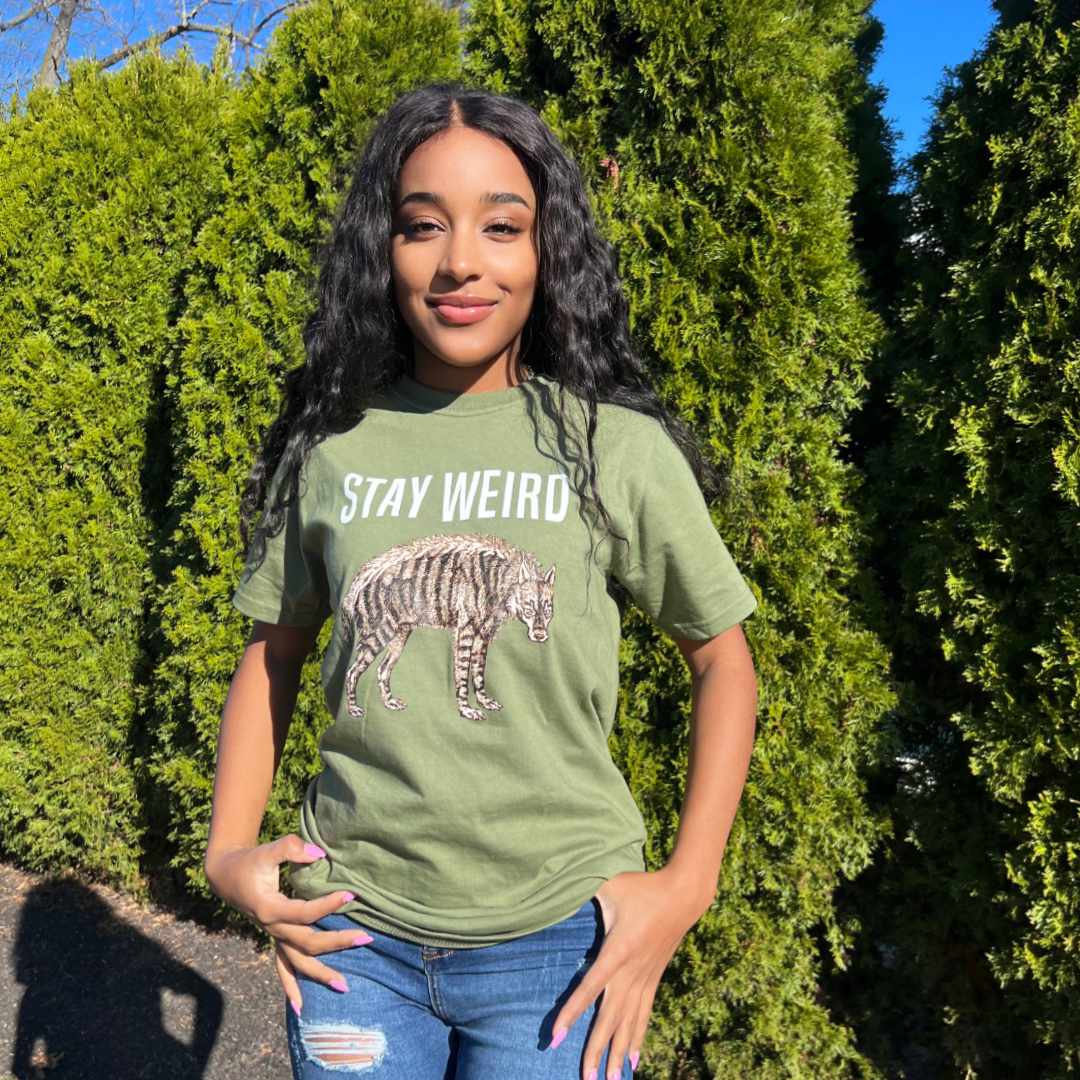This T-shirt was made for a weirdo like you!  A smiling woman models Rare Breed Organic Apparel's "Stay Weird" graphic tee in a natural outdoor setting.